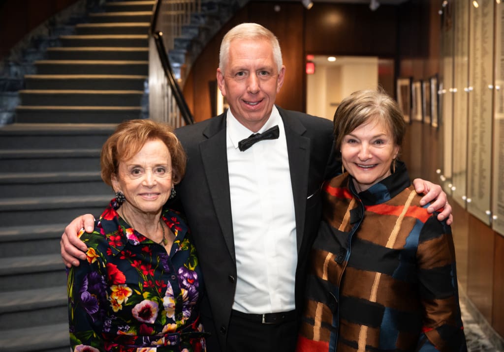 A man wearing a tuxedo stands with two women wearing brightly colored clothing.