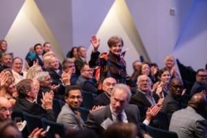 A woman in a seated audience standing up and waving.