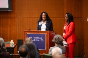 A smiling woman wearing a dark suit stands at a podium in front of an audience. A woman in a red suit stands to the right, smiling.