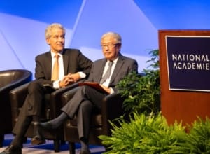 Two men wearing dark seated in chairs on a stage next to a podium with a sign that reads "National Academies."