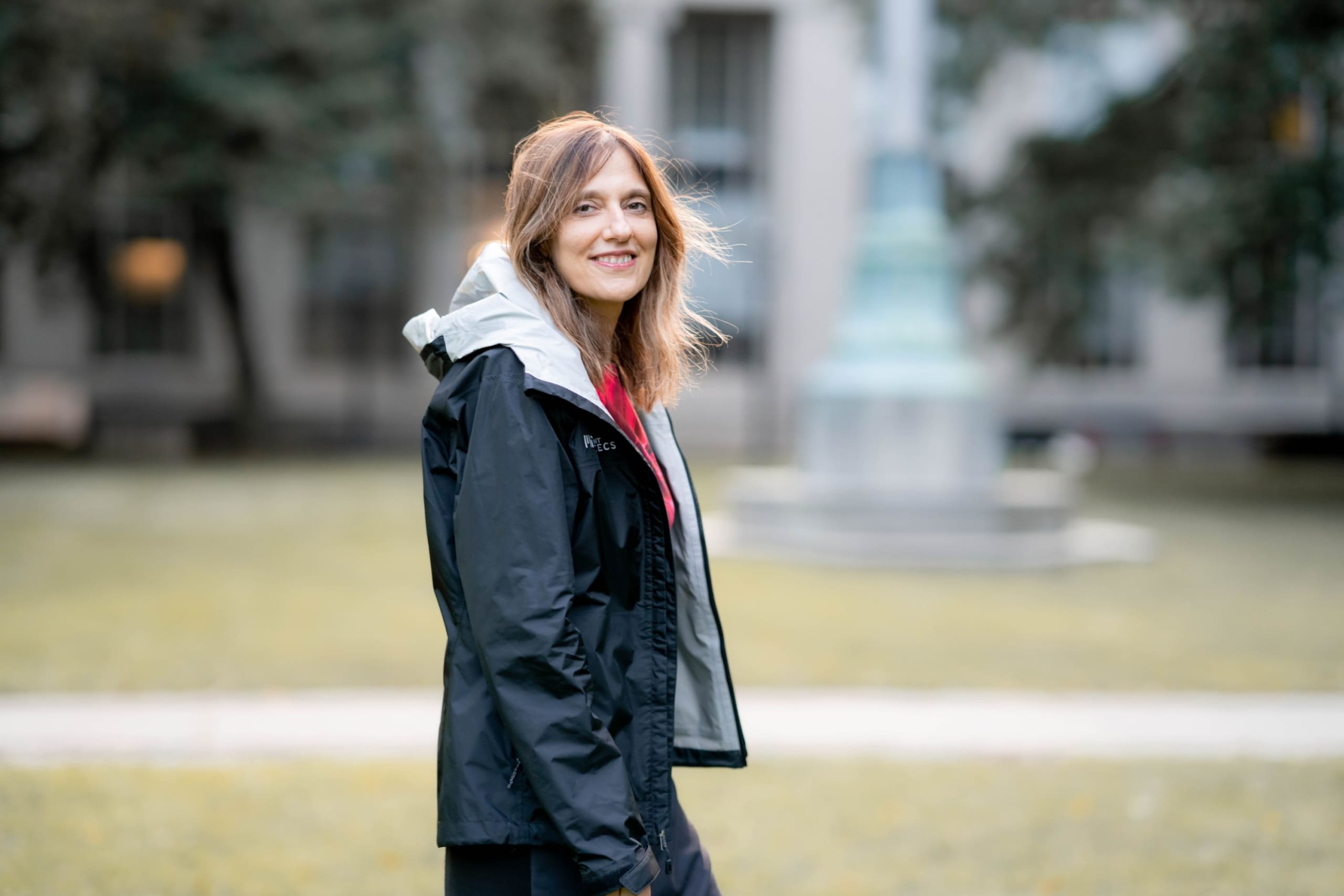 A smiling woman wearing a black hooded jacket stands on the lawn at a campus.